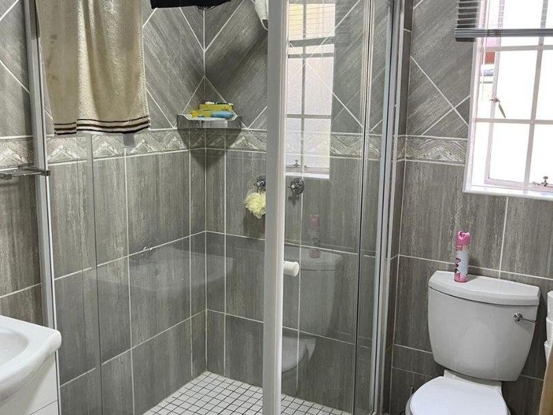 A functional bathroom featuring a corner shower enclosure, tiled walls, a standard toilet, and a vanity basin with natural light from a window.
