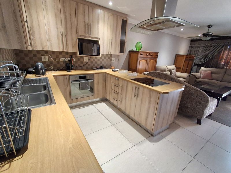 Open-plan kitchen featuring light wood cabinetry, laminate countertops, and integrated appliances, flowing seamlessly into a comfortable living area with tiled flooring throughout.