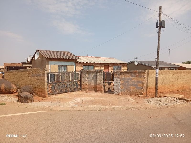 Front view of a single-story house featuring a perimeter boundary wall, metal gates, and corrugated metal roofing.
