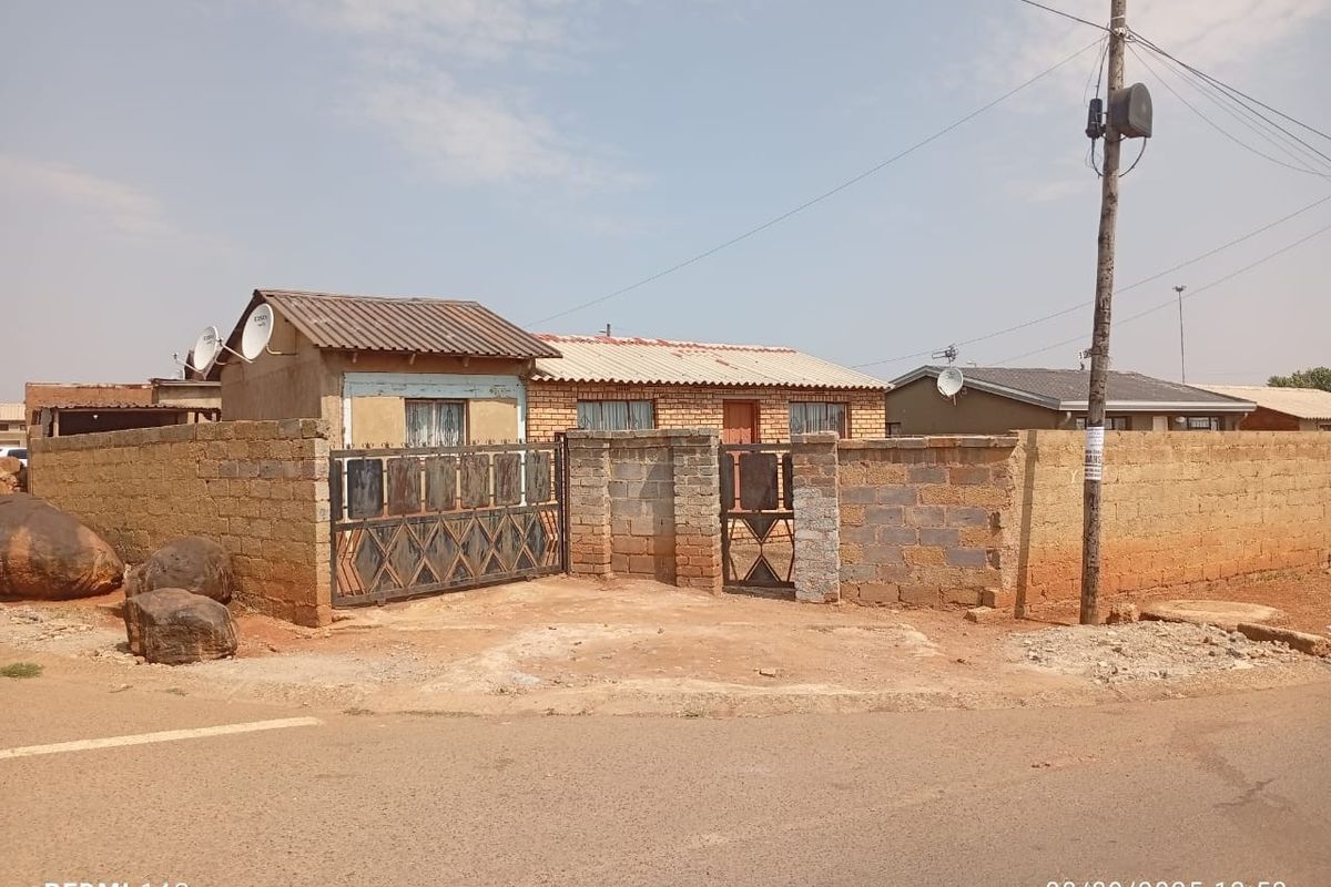 Front view of a single-story house featuring a perimeter boundary wall, metal gates, and corrugated metal roofing.