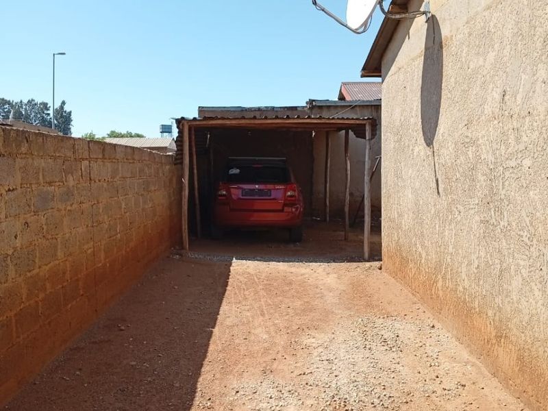 A basic outdoor carport structure with a gravel driveway and boundary wall, featuring a mounted satellite dish.