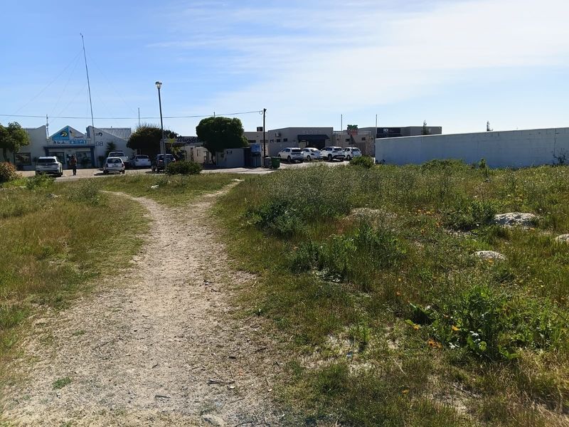 A vacant residential plot featuring an unpaved access path and boundary wall, situated in a developed area with nearby commercial properties.