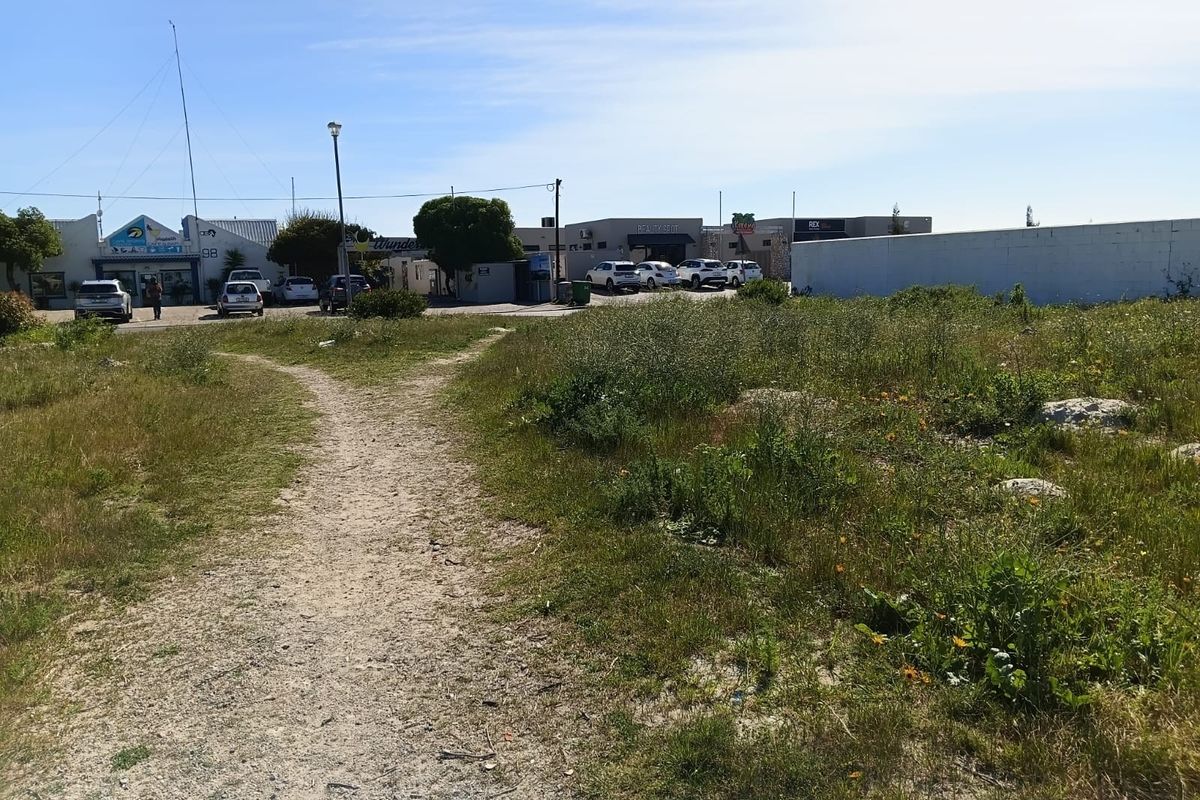 A vacant residential plot featuring an unpaved access path and boundary wall, situated in a developed area with nearby commercial properties.