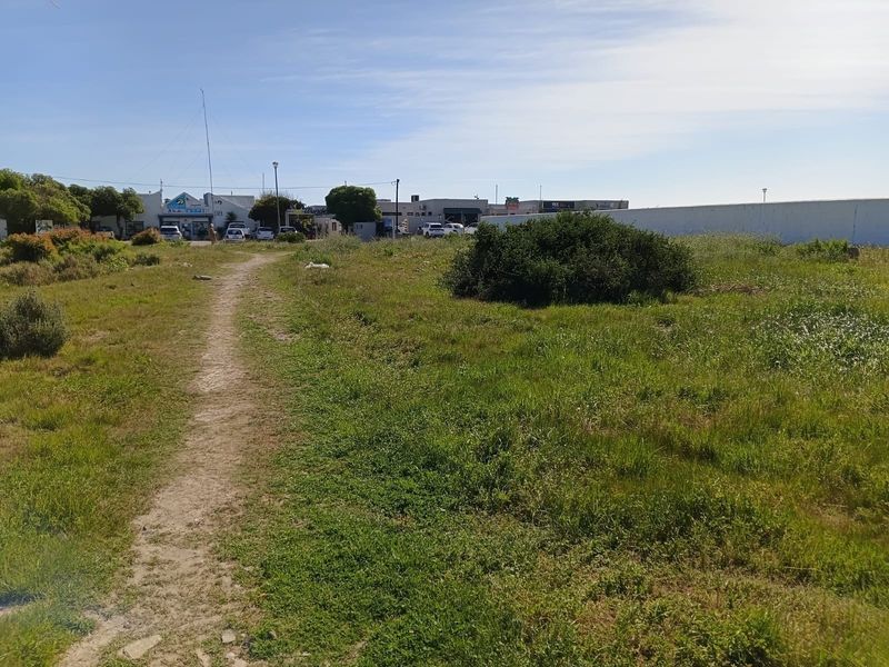 A view of an undeveloped residential plot featuring natural grassland and a dirt pathway, with surrounding commercial buildings visible in the background.