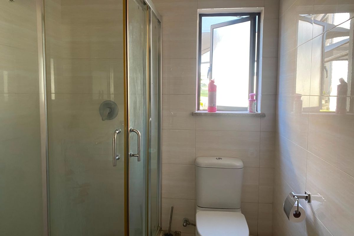 A functional bathroom featuring a glass shower enclosure, a standard toilet, and neutral wall tiling with a window for natural light.
