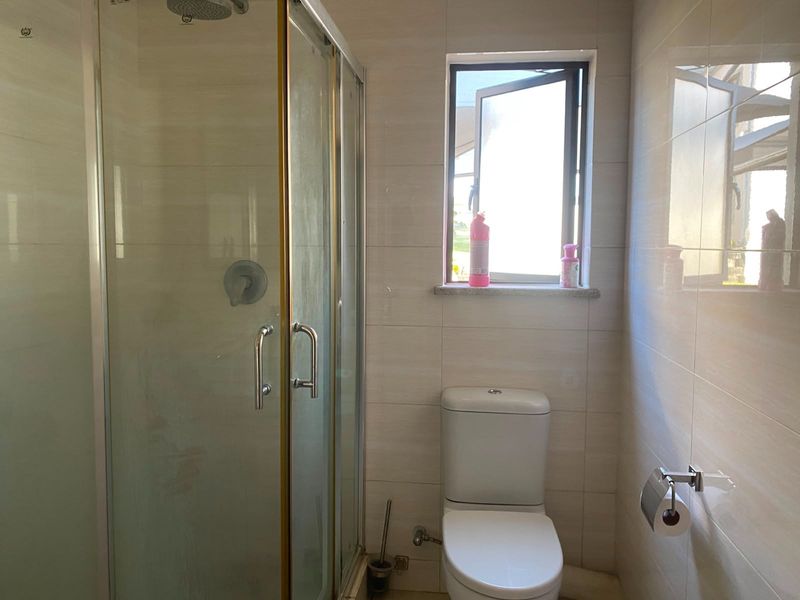 A functional bathroom featuring a glass shower enclosure, a standard toilet, and neutral wall tiling with a window for natural light.