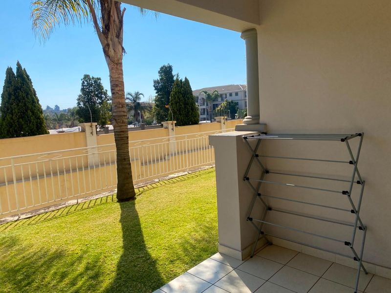 A covered patio area featuring tiled flooring and a decorative column, overlooking a private garden space with a metal perimeter fence.