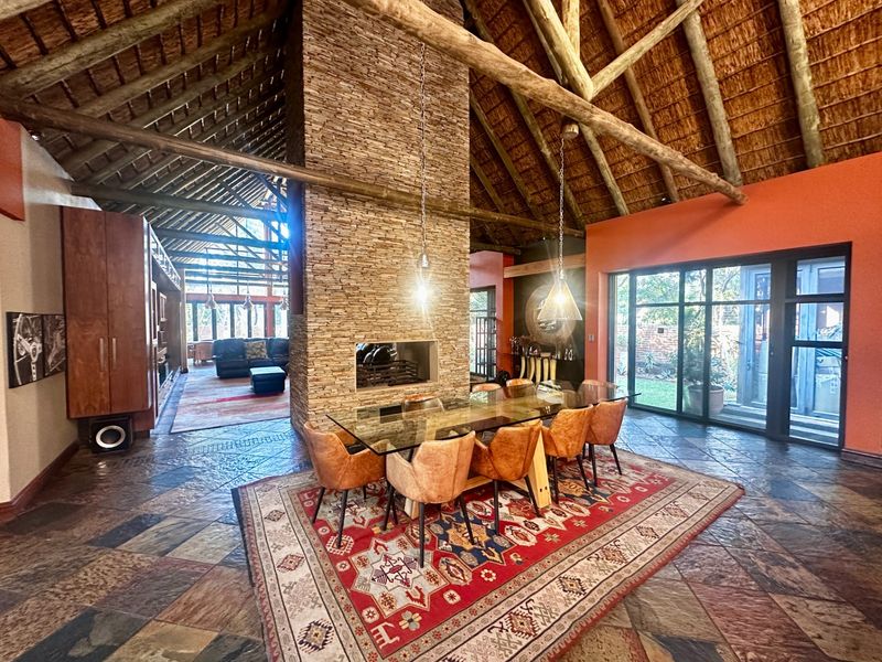 Spacious open-plan living and dining area featuring a high thatched ceiling with exposed timber beams, a central stone fireplace, and slate tile flooring.