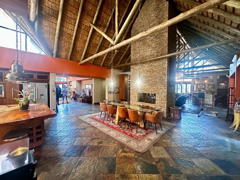 Spacious open-plan living area featuring a high thatched roof with exposed timber trusses, a central stone-clad fireplace, and durable slate tile flooring.
