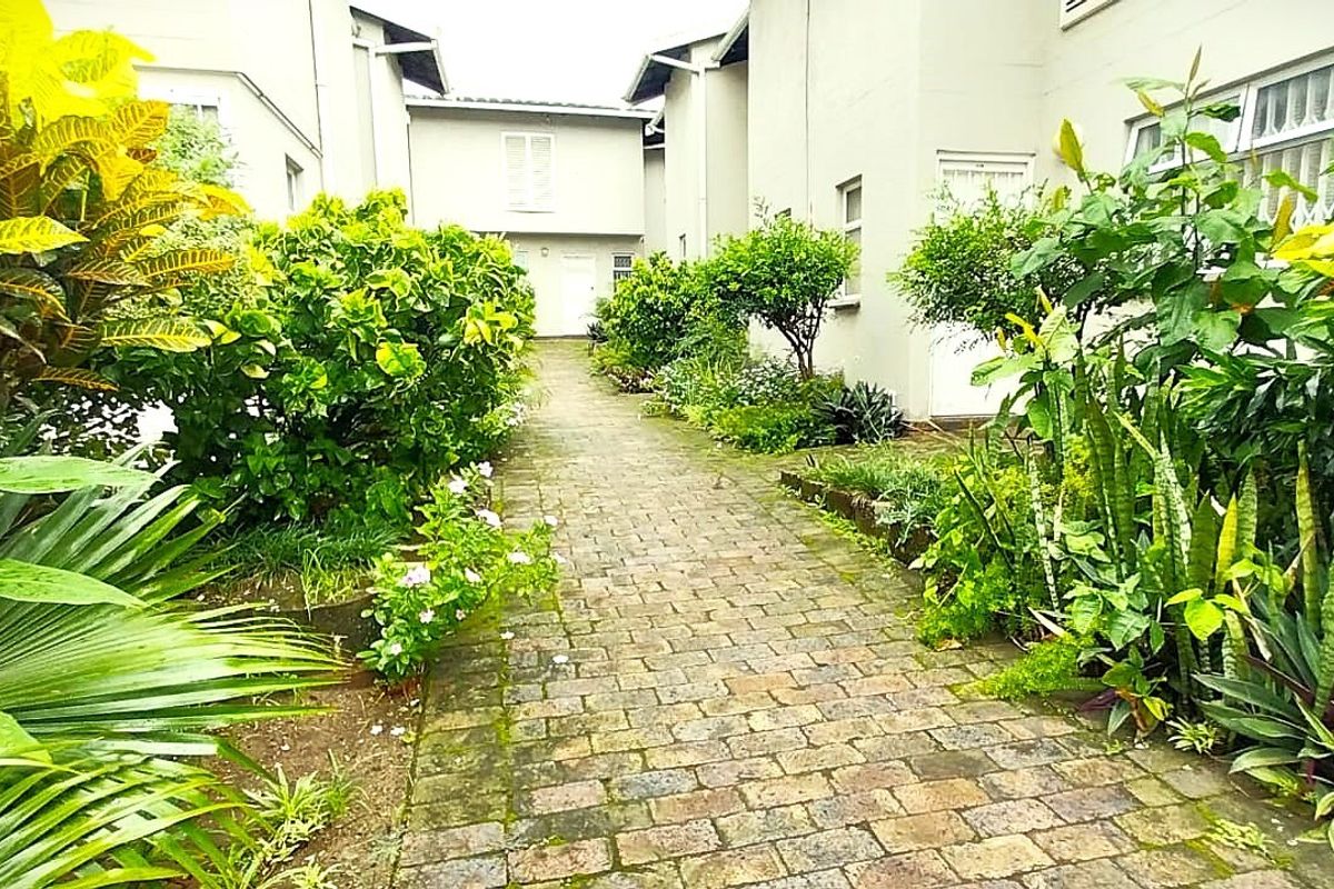 A landscaped garden pathway featuring cobblestone paving and lush greenery leading between residential building structures.