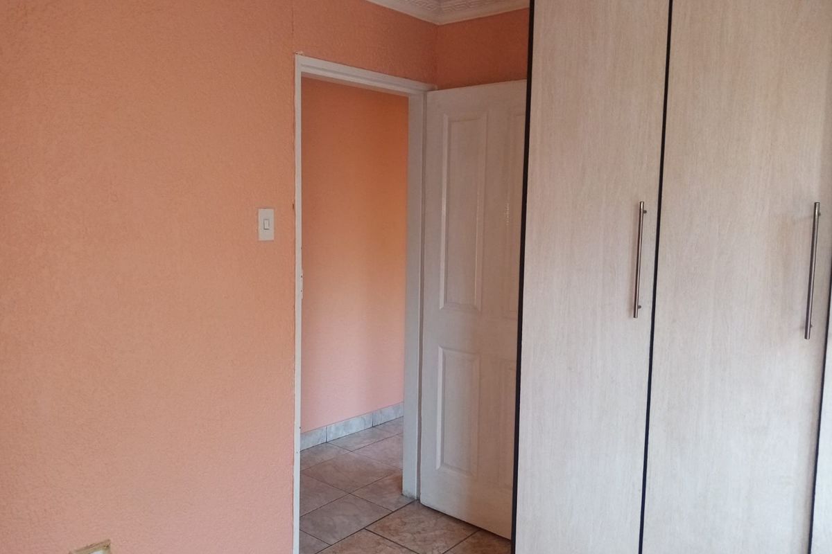 A simple bedroom featuring built-in wardrobes, tiled flooring, and peach-toned walls, leading into an adjacent hallway.