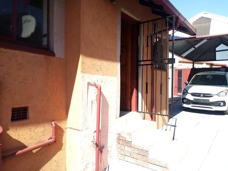Exterior view of the property entrance featuring a security gate, brick steps, and adjacent carport area with visible exterior plumbing.
