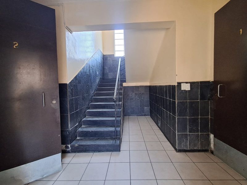 A communal hallway area featuring a staircase with a metal handrail, tiled flooring, and dark tiled wall cladding.