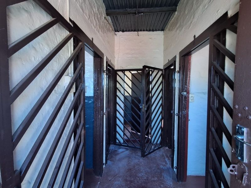 A narrow hallway featuring security gates, exposed brick walls, and a corrugated iron ceiling, showing signs of significant wear and deterioration.