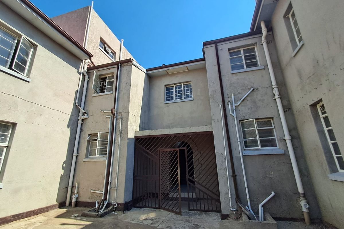 Rear exterior view of the property featuring a security gate, multiple casement windows, and exposed plumbing infrastructure against a weathered plaster facade.