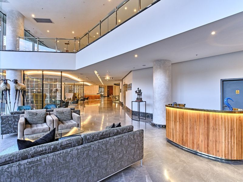 A spacious, modern apartment building lobby featuring marble flooring, a curved wooden reception desk, and a mezzanine level with glass balustrades.