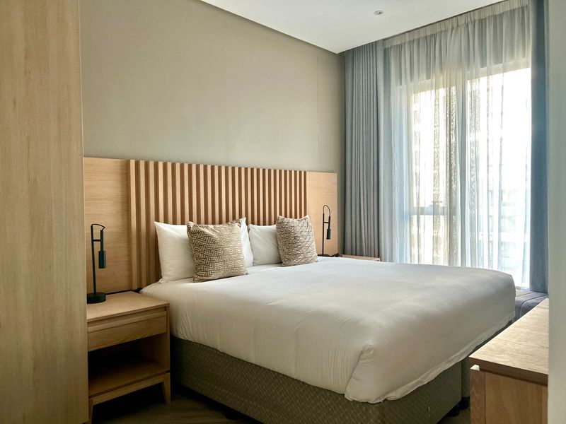 A modern, well-maintained bedroom featuring a custom timber-slat headboard, integrated cabinetry, and floor-to-ceiling curtains allowing for natural light.