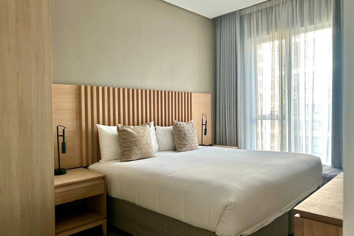 A modern, well-maintained bedroom featuring a custom timber-slat headboard, integrated cabinetry, and floor-to-ceiling curtains allowing for natural light.