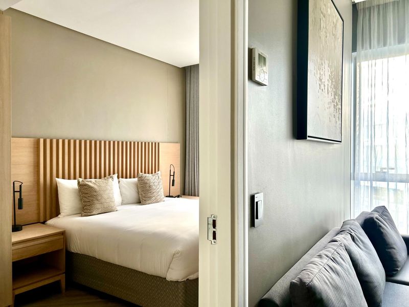 A modern bedroom featuring a contemporary slatted timber headboard, neutral wall tones, and large windows with sheer curtains, viewed through a doorway.
