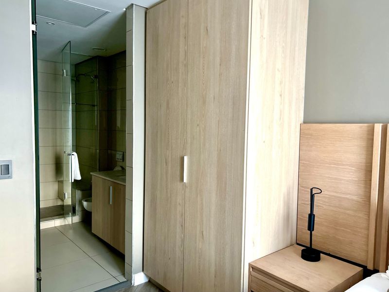 A modern bedroom featuring a light wood built-in wardrobe and an adjoining en-suite bathroom with a glass-enclosed shower and vanity.