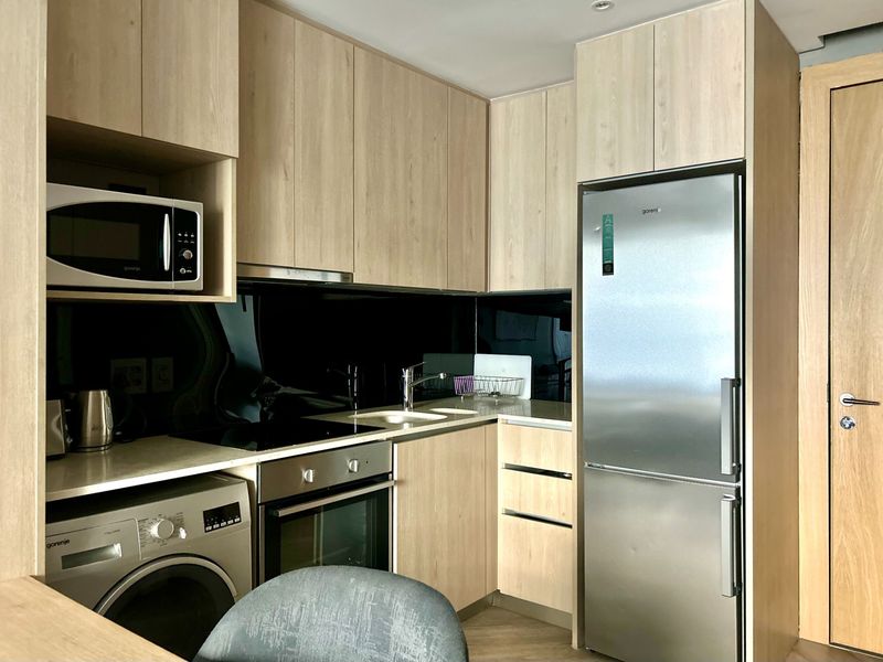 Modern compact kitchen featuring light wood cabinetry, integrated appliances, stone countertops, and a sleek black glass splashback.