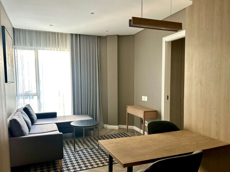 A modern, well-lit living area featuring laminate flooring, neutral wall tones, and floor-to-ceiling curtains leading to a balcony.