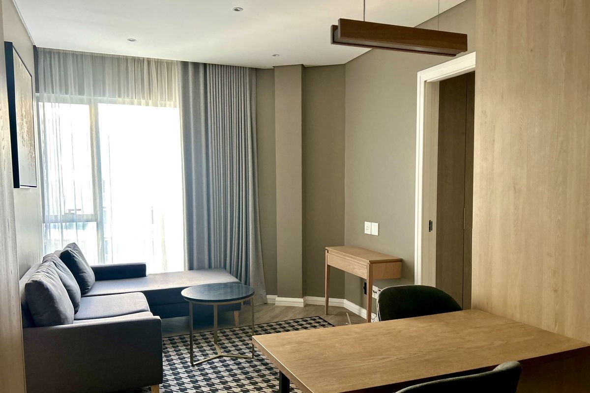 A modern, well-lit living area featuring laminate flooring, neutral wall tones, and floor-to-ceiling curtains leading to a balcony.