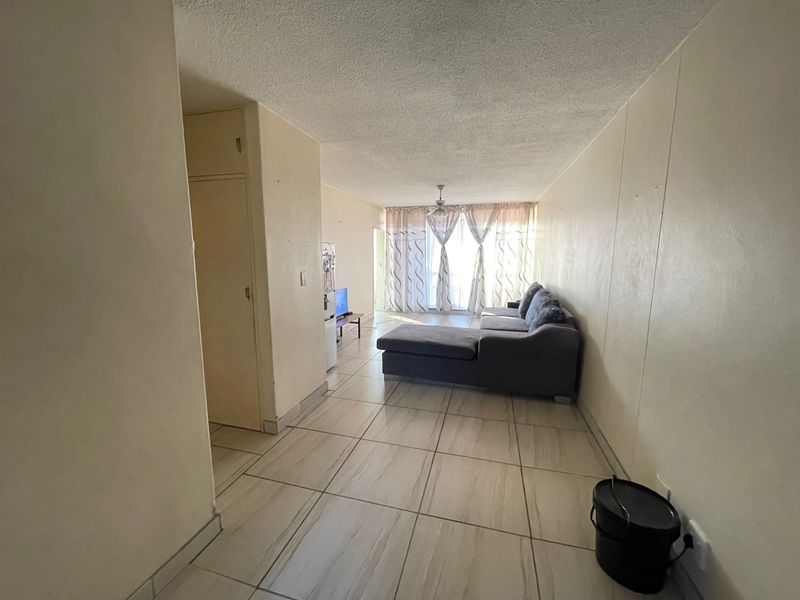 A living room area featuring tiled flooring, a ceiling fan, and a built-in storage cupboard, leading towards a window with curtains.