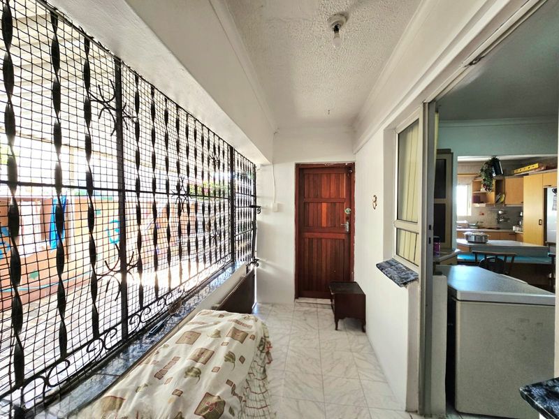 A covered patio or enclosed walkway featuring tiled flooring, a timber door, and extensive security screening, providing access to the kitchen area.