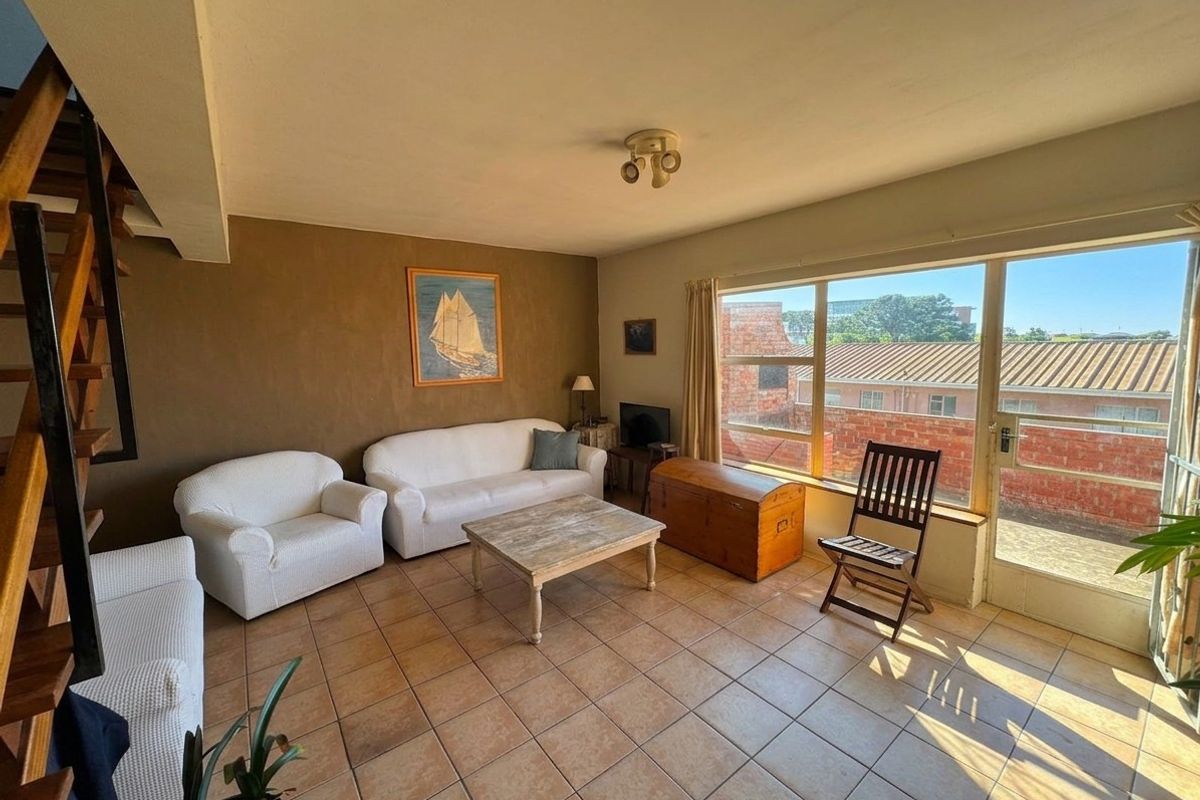 A living room featuring tiled flooring, a wooden staircase, and large windows providing natural light and access to an outdoor area.
