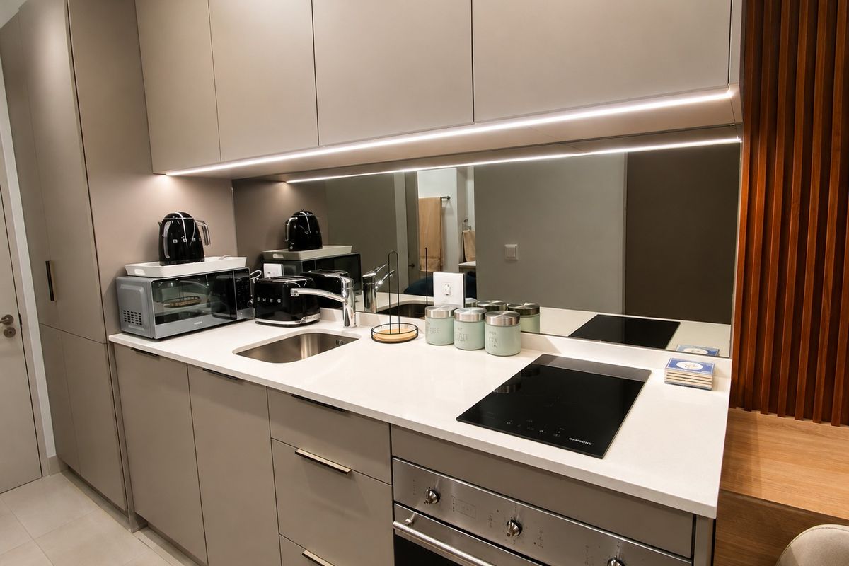 Modern, compact kitchen featuring sleek handleless cabinetry, stone countertops, a mirror splashback, integrated hob, and built-in oven with ambient under-cabinet lighting.