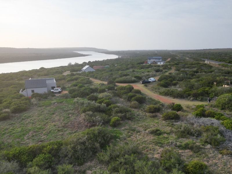 An aerial view of a coastal residential plot featuring natural vegetation, river frontage, and existing residential structures in the distance.