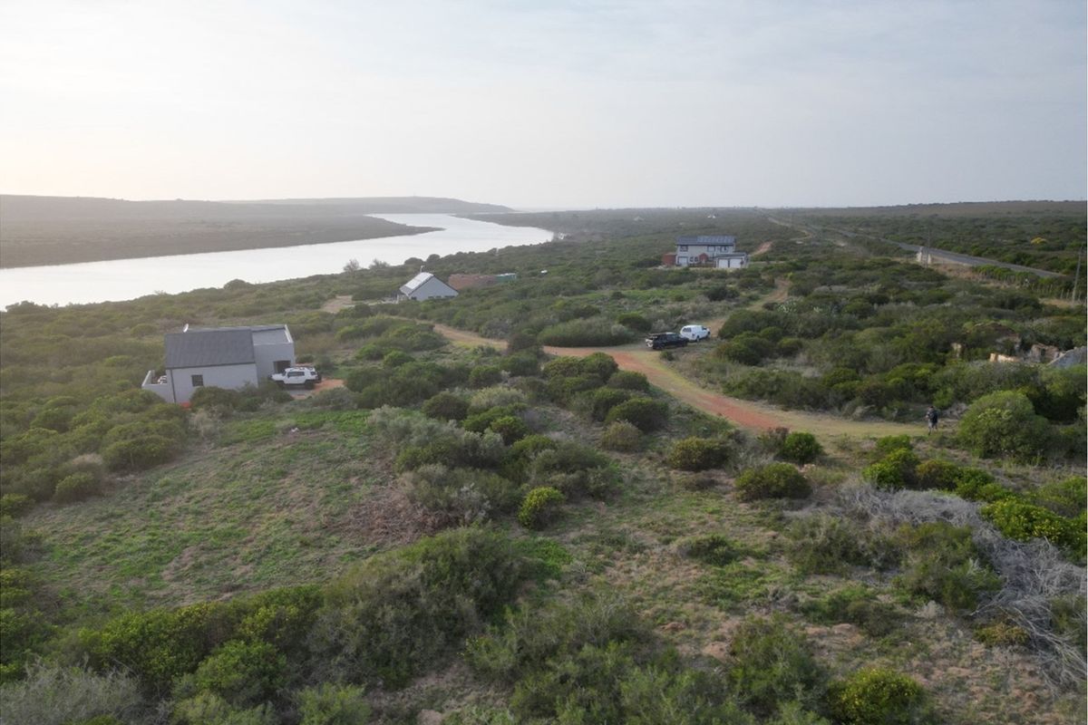 An aerial view of a coastal residential plot featuring natural vegetation, river frontage, and existing residential structures in the distance.