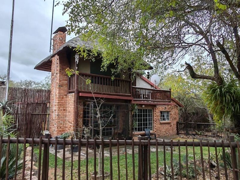 Rear view of a two-story brick house featuring a thatched roof, a wooden balcony, and secure steel window bars, set within a natural garden.
