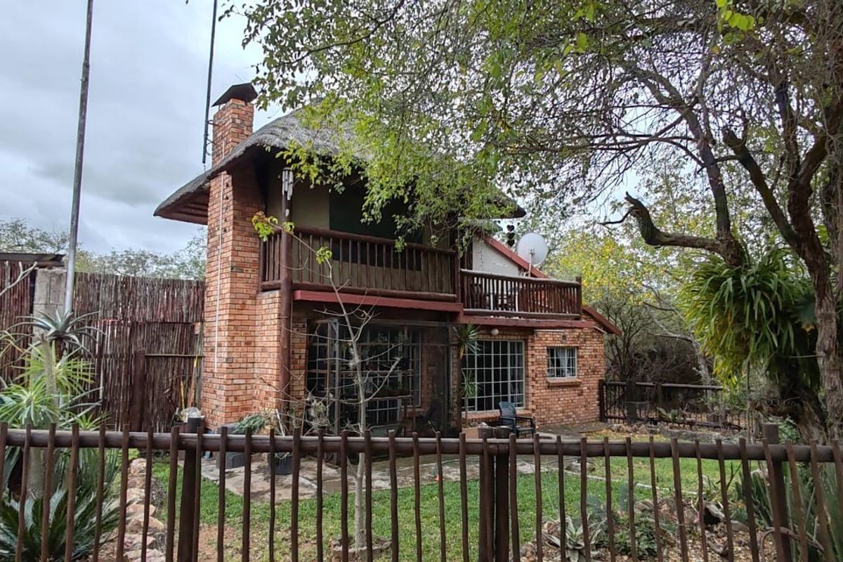 Rear view of a two-story brick house featuring a thatched roof, a wooden balcony, and secure steel window bars, set within a natural garden.