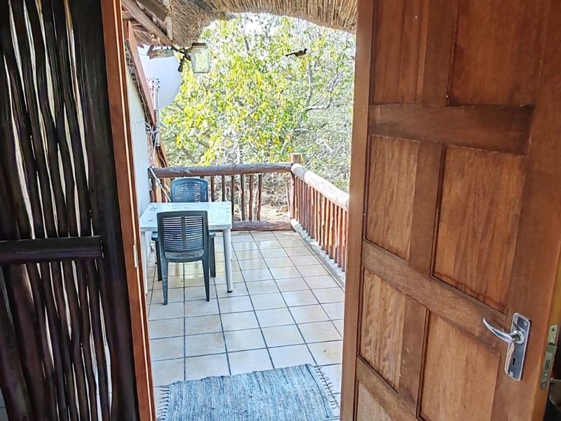 A covered outdoor patio area featuring tiled flooring, a wooden balustrade, and a thatched roof, providing a rustic transition to the garden.