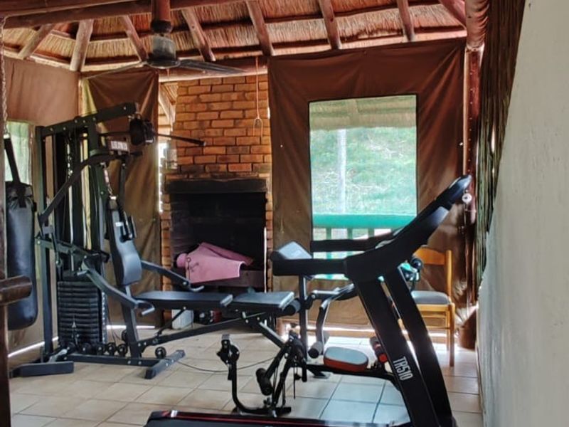 A rustic gym space featuring a traditional thatched roof with exposed timber rafters, tiled flooring, and a built-in brick fireplace.