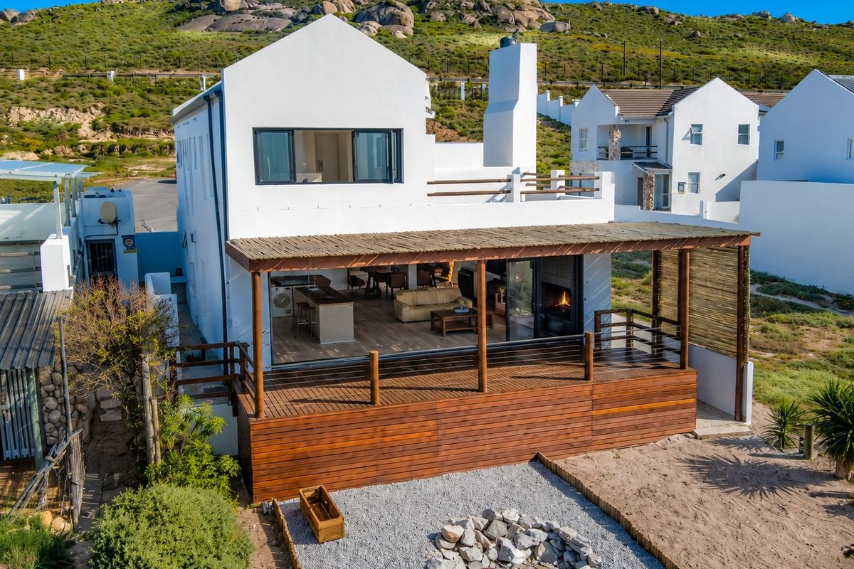Aerial view of the rear elevation featuring a spacious timber deck, pergola, outdoor fireplace, and a gravel fire pit area.