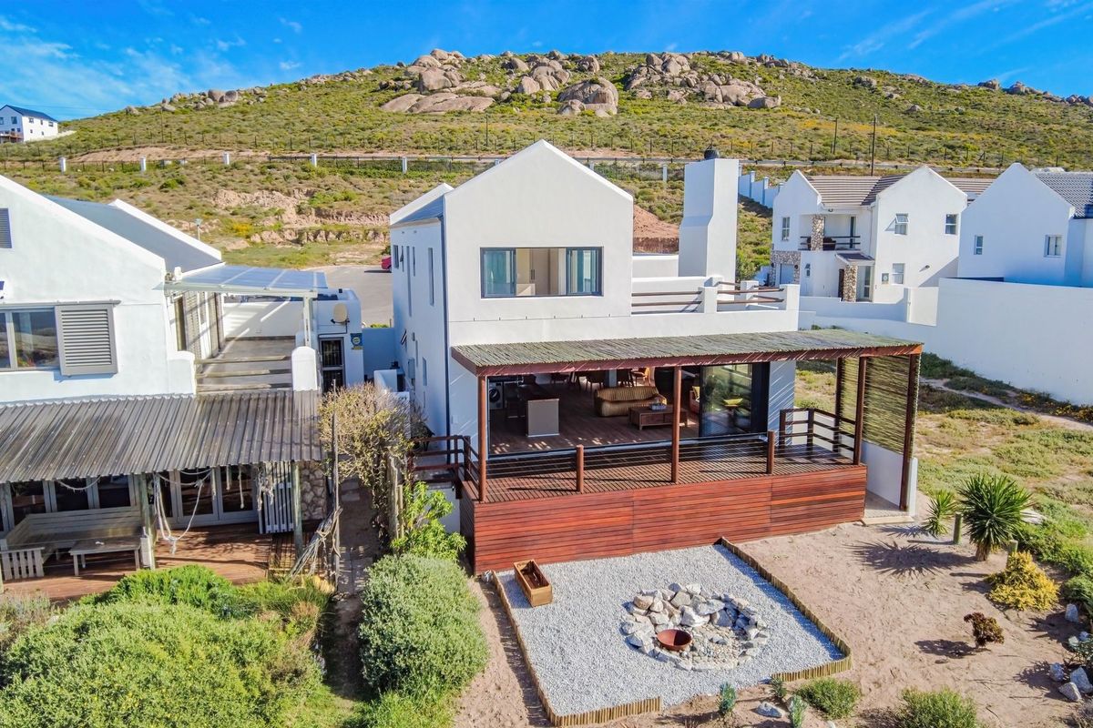 Aerial view of a modern coastal home featuring a large timber deck, covered patio area, and a landscaped fire pit in the rear garden.