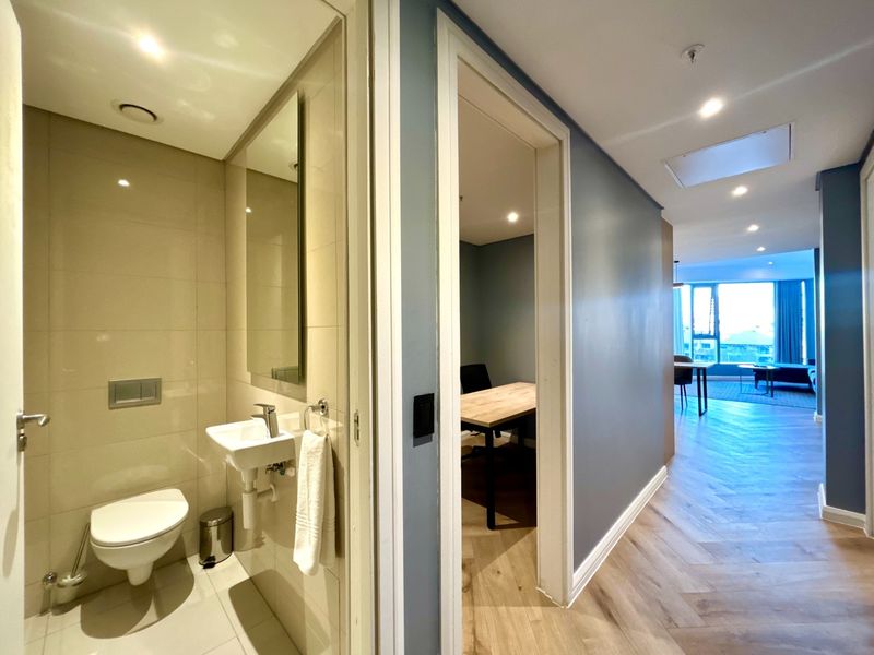 A modern hallway featuring herringbone timber flooring, recessed lighting, and access to a compact guest bathroom with wall-mounted fixtures.
