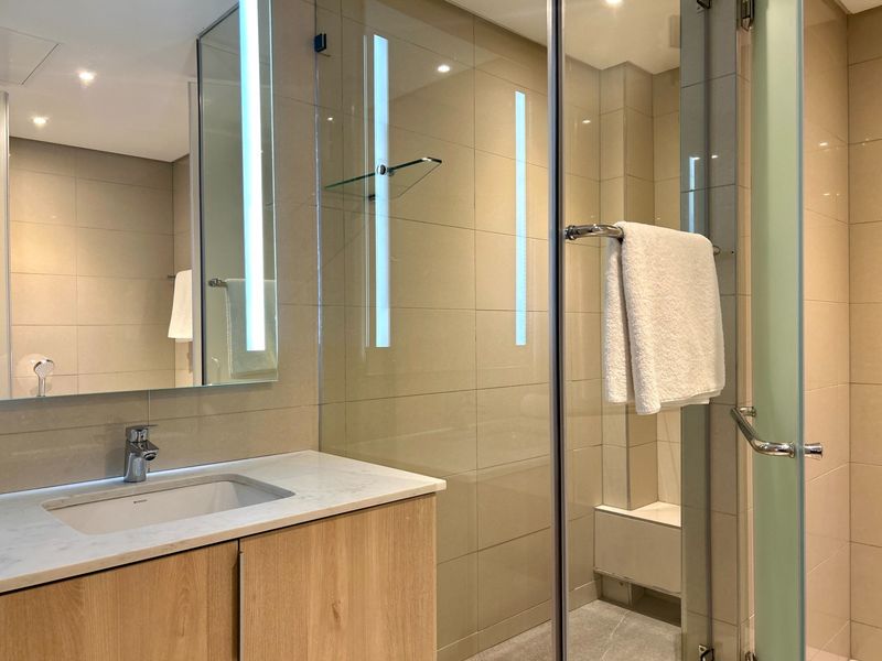 Modern bathroom featuring a stone-topped vanity, integrated mirror lighting, and a spacious glass-enclosed shower with neutral wall tiling.