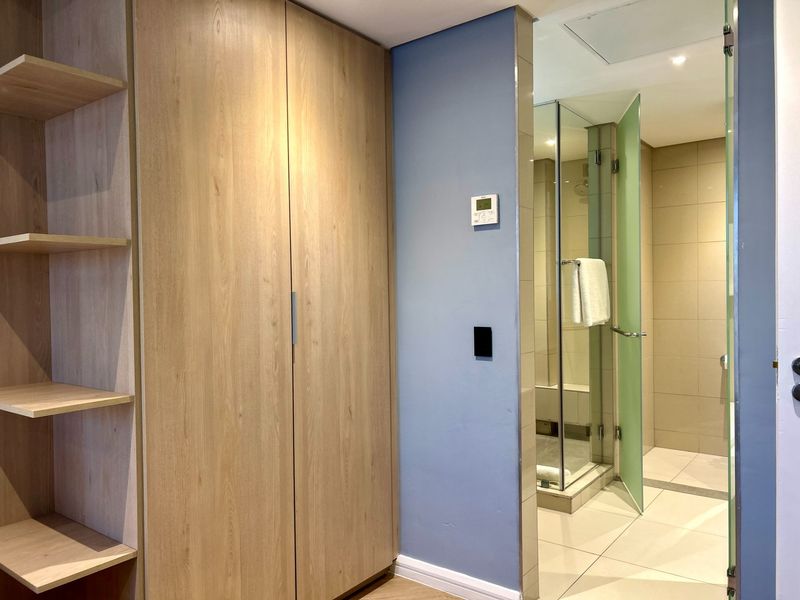 A modern bedroom area featuring built-in wooden cabinetry with open shelving and an adjacent en-suite bathroom with a glass-enclosed shower.