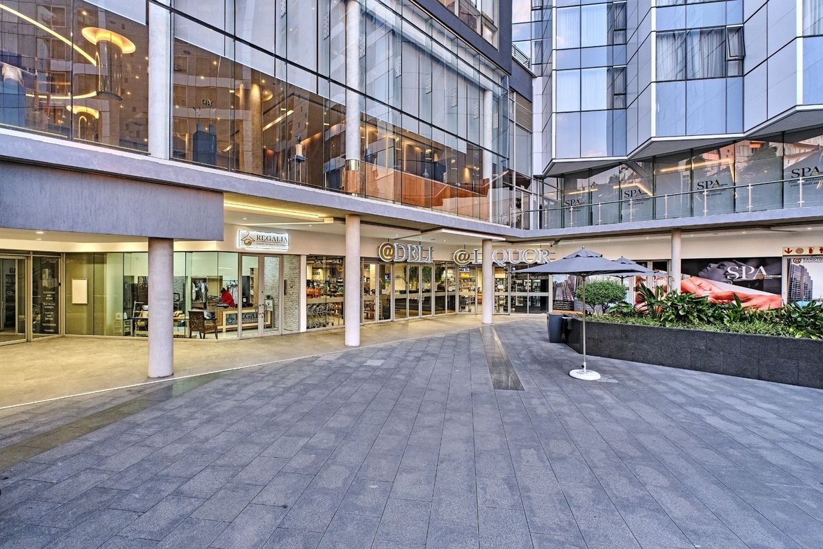 Modern commercial courtyard area featuring glass facade architecture, paved pedestrian space, and integrated retail storefronts within a contemporary apartment complex.