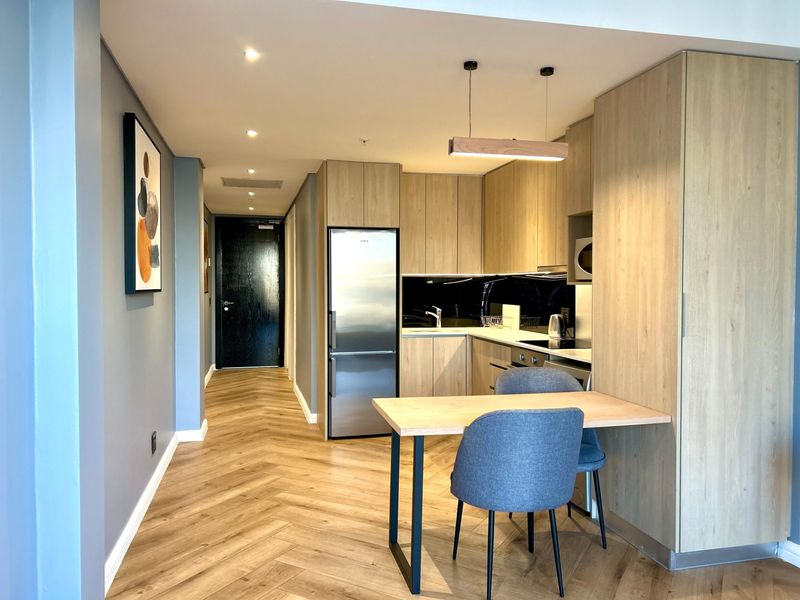 Modern open-plan kitchen featuring light wood cabinetry, herringbone flooring, integrated appliances, and recessed lighting, seamlessly flowing into the apartment entrance area.