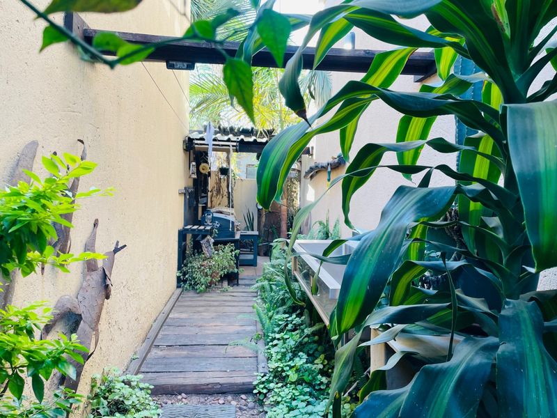 A narrow outdoor patio area featuring wooden decking, a built-in sink, and a pergola structure, surrounded by lush garden greenery.