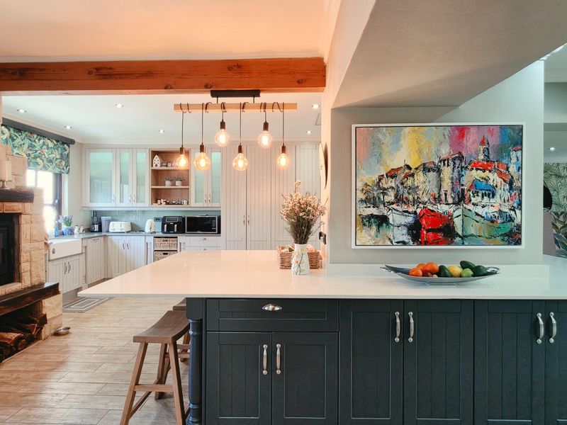 Spacious open-plan kitchen featuring a large island, shaker cabinetry, farmhouse sink, and a cozy stone fireplace with an exposed timber beam.