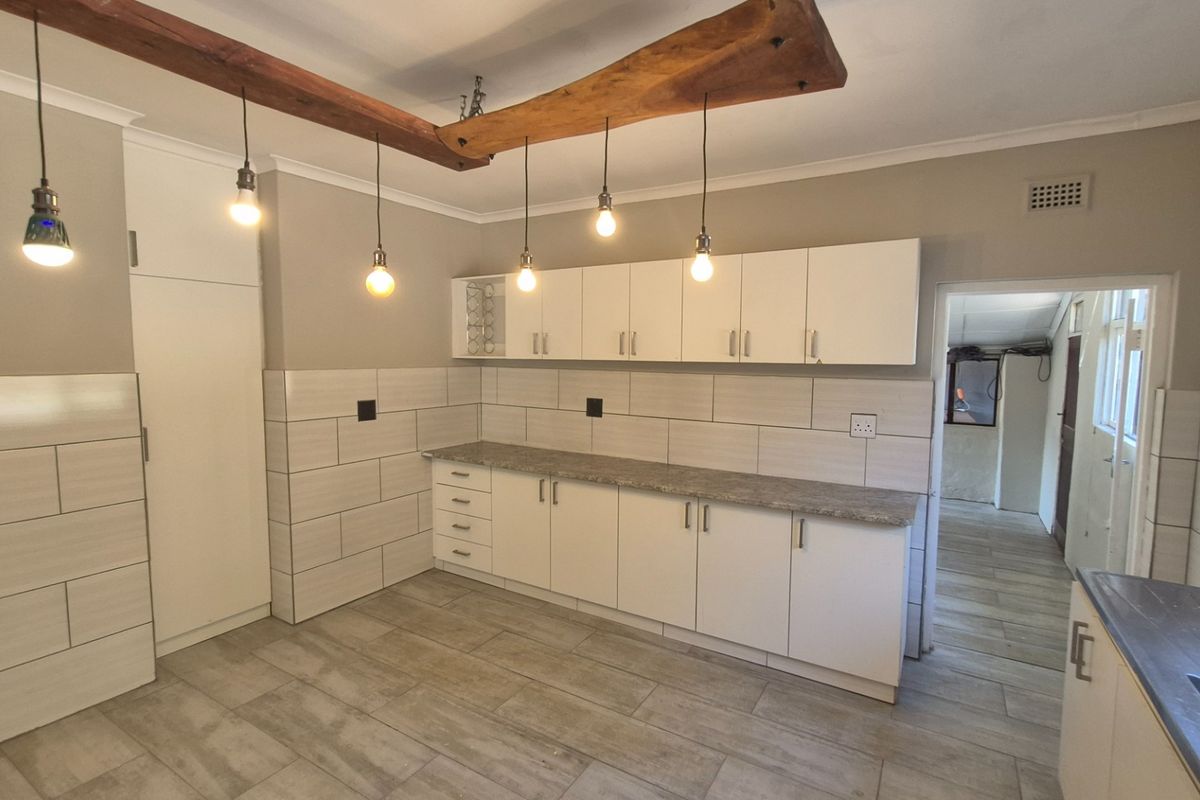A modern, clean kitchen featuring white cabinetry, stone-look laminate countertops, tiled backsplash, and stylish pendant lighting suspended from a rustic timber beam.