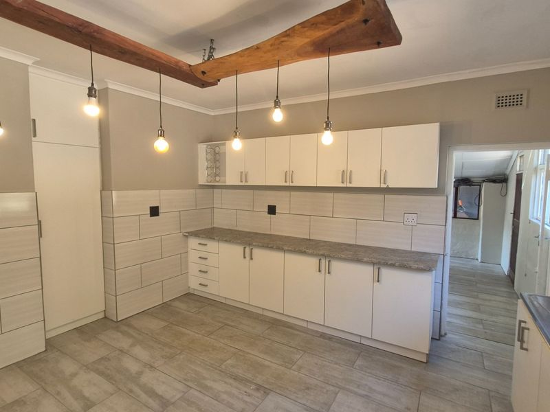 A modern, clean kitchen featuring white cabinetry, stone-look laminate countertops, tiled backsplash, and stylish pendant lighting suspended from a rustic timber beam.