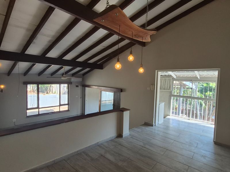 Spacious living area featuring vaulted ceilings with exposed timber beams, tiled flooring, and large windows providing natural light and access to an outdoor area.