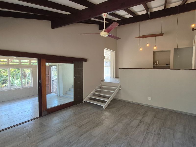 Spacious living area featuring exposed timber ceiling beams, tiled flooring, and a unique staircase, offering a versatile open-plan layout with natural light.